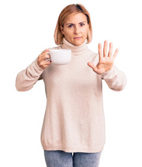 Young blonde woman holding coffee with open hand doing stop sign with serious and confident expression, defense gesture