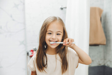 Portrait of a girl with Down syndrome brushing her teeth. Happy little girl brushing her teeth in front of a bathroom mirror. Morning hygiene.