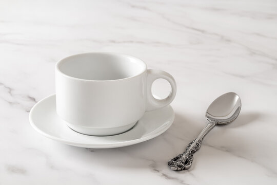 White cup on saucer and teaspoon over marble table. Empty clean porcelain crockery for drink design mockup. Tableware, breakfast, tea and coffee concept. High key image.
