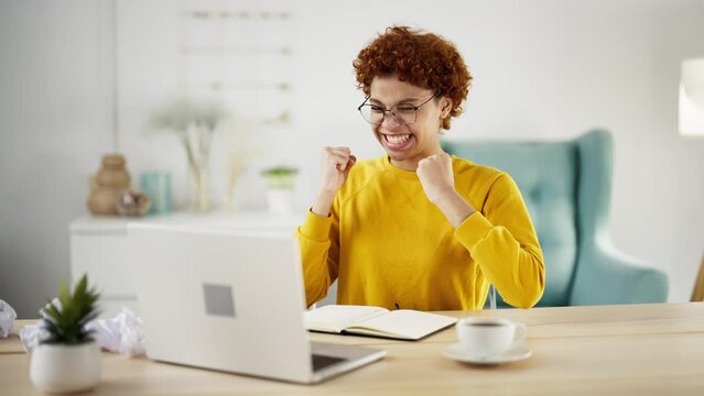 Overjoyed amazed african american woman rejoicing achieving goal, showing win victory hands gesture shaking fists over head looking at laptop screen sitting at table. Girl winner celebrates success.