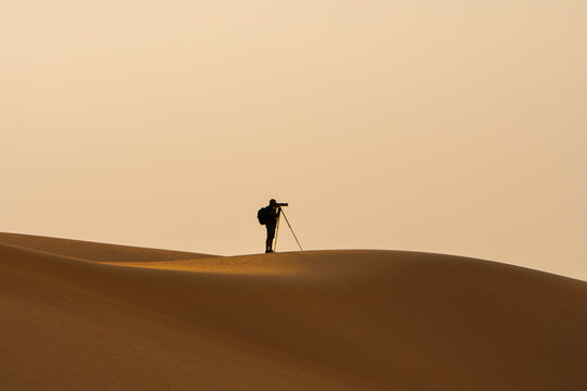 A man settting up his camera gear for photography at dawn on a sand dune in the empty quarter of the United Arab Emirates. Orange sand, hazy sky.