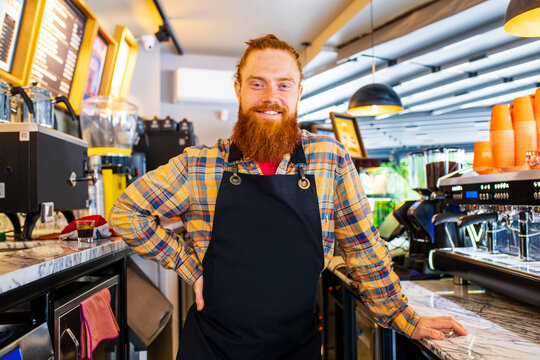 Professional barista young redhaired ginger bearded man in black apron working in coffee shop