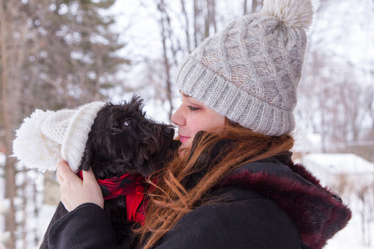 Medium shot of cute red-haired young woman in winter clothes holding her miniature black Schnauzer with wooly hat staring at her lovingly