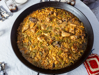 Beautiful overhead view of a typical Valencian paella, with the original ingredients chicken, rabbit, green and white beans. Table with glasses, plates, cutlery and napkins. Exquisite homemade paella