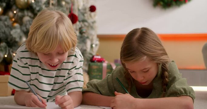 boy and girl write together letters with wish list to santa claus using color pencils laying on floor near christmas tree.