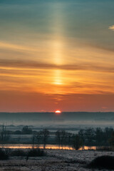 Fototapeta premium Sunrise over a river valley with trees and meadows on a foggy morning. A light pillar is an atmospheric optical phenomenon, a vertical beam of light from the Sun. Landscape of late autumn or winter