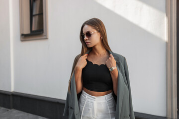 Elegant beautiful young business woman with vintage sunglasses in fashionable urban clothes with a green blazer stands near a white wall on the street