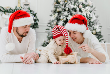 Fototapeta na wymiar Happy family under Christmas tree. baby boy in Santa Claus hat with gifts under Christmas tree with many gift boxes presents. Happy Holidays, New year. Cozy warm winter evening at home. Xmas time