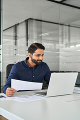 Busy Indian business man holding financial report looking at laptop calculating assessments analyzing project overview, male company hr using computer working online in office, vertical shot.