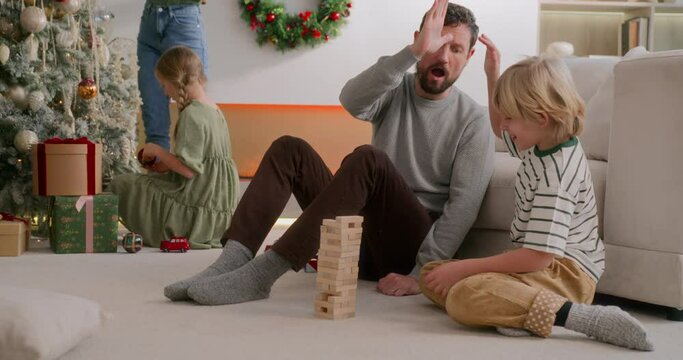 dad and son play jenga together on floor at home, smiling and laughing, mom and daughter decorate christmas tree on background