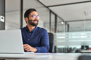 Happy Indian business man ceo executive looking away working on laptop. Smiling professional company manager or employee thinking of future financial success and growth at work in office. Copy space.