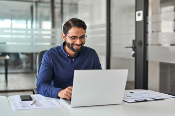 Happy Indian business man looking at laptop using computer working in office. Smiling busy professional businessman employee or company executive cheerful about online project goals achievement. - obrazy, fototapety, plakaty