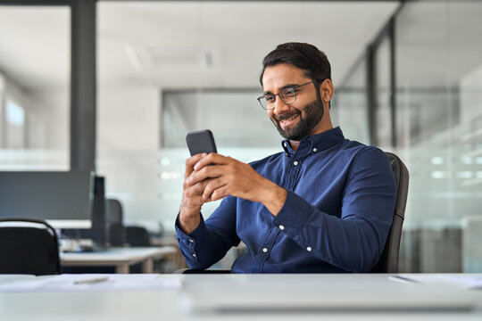 Happy busy Indian business man executive using mobile phone working in office. Professional businessman employee looking at smartphone checking banking apps typing on cell sitting at workplace desk. - Powered by Adobe