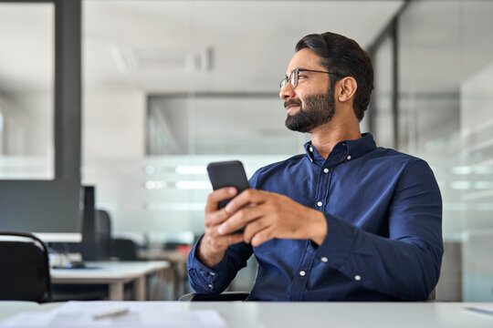 Smiling professional Indian businessman employee looking aside holding smartphone sitting at desk. Happy busy business man executive using cell phone thinking of new mobile solutions working in office