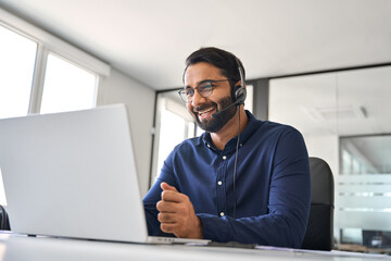 Smiling contract service telemarketing operator using laptop having conversation. Happy Indian call center agent wearing headset talking to client working in customer support office. Authentic shot