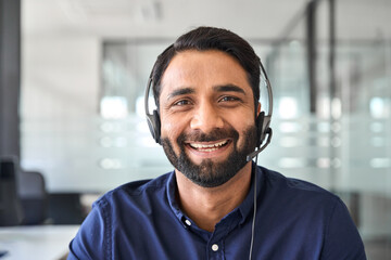 Male Indian contract service representative telemarketing operator smiling to camera. Happy man call center agent or salesman wearing headset working in customer support office. Close up portrait.