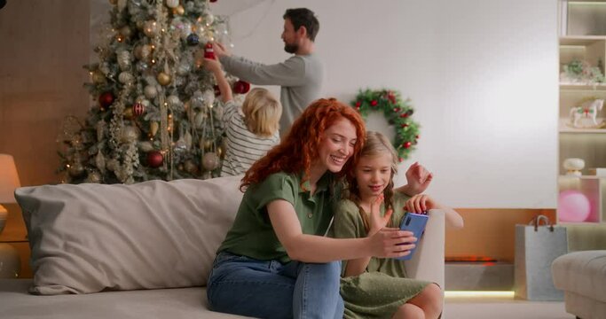 mom and girl make video call to far distance relatives on holidays to wish merry christmas. dad and son decorate christmas tree on background and wave hello