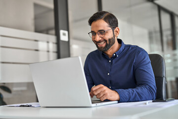 Happy professional Indian business man company employee, male worker, manager or financial analyst smiling looking at computer satisfied with work results and profit growth sitting at office desk.