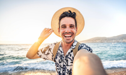 Happy handsome man taking selfie pic with cellphone outside - Male tourist enjoying summer vacation at beach holiday - Travel life style concept with smiling guy laughing at camera - obrazy, fototapety, plakaty
