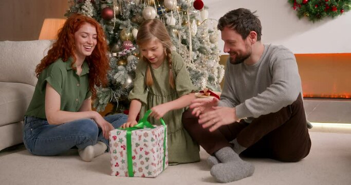 mom, dad and daughter sit together on floor in living room near decorated christmas tree, boy unwrap his present. feel happy, joyful and inspired, smiling and laughing