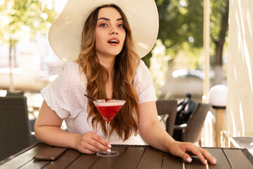 Pretty modern woman wear straw hat holding strawberry cocktail in glass sitting on summer bar