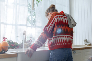 young woman in red sweater stays in kitchen leaning on sink feeling upset. thoughtful and...
