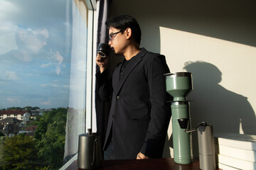 asian man wear black suit dark shade glasses stand in fornt of desk books make confidence mood and coffee with city window view and shadow on wall