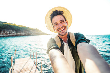 Happy handsome man taking selfie pic with cellphone outside - Male tourist enjoying summer vacation at beach holiday - Travel life style concept with smiling guy laughing at camera - obrazy, fototapety, plakaty