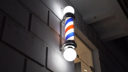 Barber shop spiral light pole with rotating blue and red stripes, shining at night while hanging on the tiled exterior wall by the barber shop entrance door. - Powered by Adobe