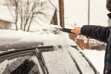 A young man removes snow from the roof and car windows with a brush. Heavy snowfall and cyclone.