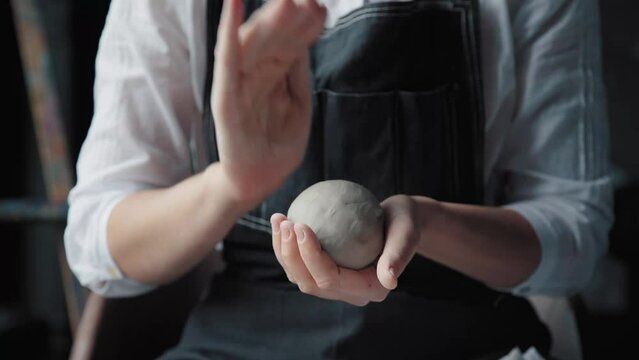 A woman in a black apron holds white clay in hands, makes a ball out of it, and warms it in palms. Medium shot, only the hands in the frame.