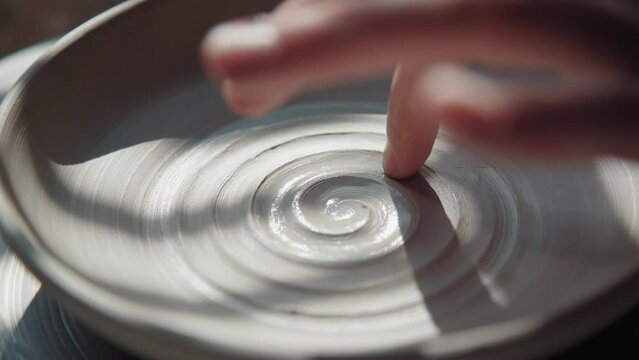 Potter female makes a plate from white clay and draws a spiral inside, using a finger. Process: the ceramist makes a pattern inside the pottery of clay, making a spiral. Close-up, only hands in frame