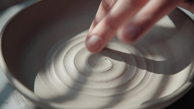 Potter female makes a plate from white clay and draws a spiral inside, using a finger. Process: the ceramist makes a pattern inside the pottery of clay, making a spiral. Close-up, only hands in frame