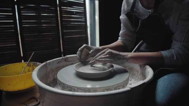 Woman works on a potter's wheel in a workshop with dim lights. She sits in an apron in front of the wheel and sponge off the extra clay. The wheel rotates with the product in the center.