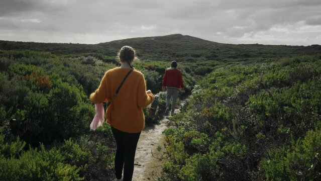 Rear view of couple strolls apart along the green bushy area, enjoying the calming native environment of Western Australia. Young adult female in her vibrant orange sweater and her partner walks on
