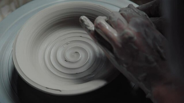 The clay product rotates on a potter's wheel: a plate with a pattern - a spiral. In the frame potter's wheel and the hands of the master. Top view, pottery.