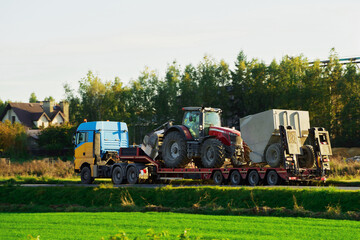 Obraz premium A heavy truck with a flatbed trailer transports a big tractor on the road. The photo shows the industrial machinery and equipment used for farming and agriculture. The truck is driving in the evening