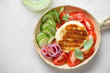 Serving pan with whole grilled halloumi and fresh vegetables on a light-beige stone background, horizontal shot, elevated view