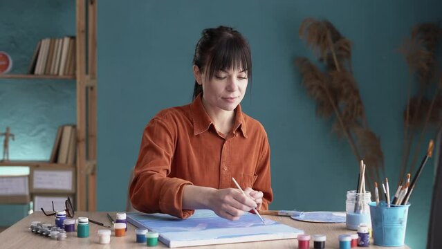 Female artist working in art studio sitting at the table with other creative tools and canvas in front of her