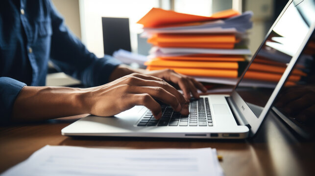 Close-up of a man's hands typing on a laptop keyboard, with a stack of paperwork beside them
