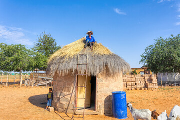 african woman climbed to repair a thatched roof on a hut, child and livestock in the yard