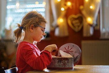 Happy little girl eating gingerbread Christmas cookies in domestic kitchen decorated with lights....