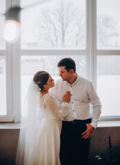 Newlyweds standing in the front of a bright window. Winter