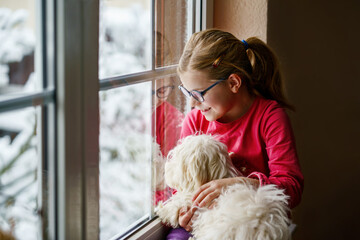 Little girl sitting by window with her pet dog Maltese at home. Happy child and cute puppy looking...