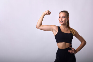 Slender young girl with a smile in sweatpants and sports top stands on gray background, showing off her biceps. Strong athletic female fitness instructor in black tracksuit poses on white background