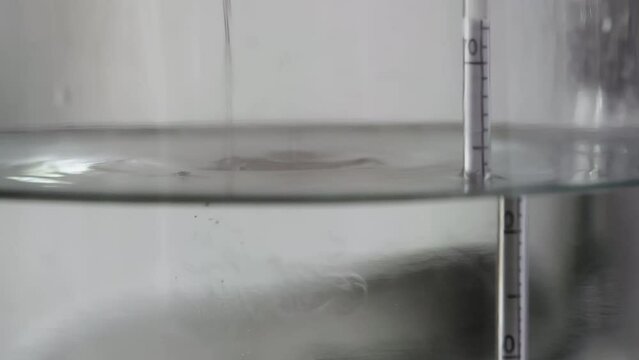 alcohol distilling process closeup - liquid distilled transparent liquid flowing in large glass jar with floating alcoholometer in it.
