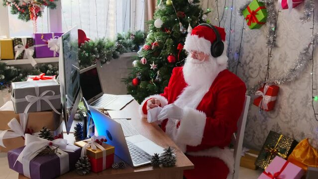 Operator in Santa Claus costume and headphones sits in office behind devices, laptops, and computer against backdrop of Xmas decorations, flickering lights of Christmas tree and many packed gift boxes