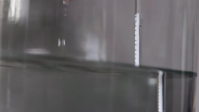 alcohol distilling process closeup - liquid distilled transparent liquid flowing in large glass jar with floating alcoholometer in it.