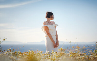 Beautiful Asian young woman in white dress outdoor in flower field under Rhodes city and above sea during sunset. embracing fresh air and engaging in outdoor activities. strong wind - element of