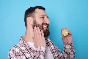 Handsome bearded man holding beard balm. Bearded man applying wax and oil to his beard on blue background.  - obrazy, fototapety, plakaty